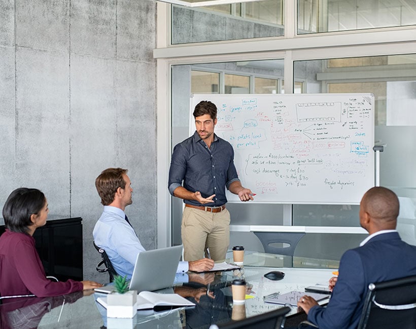 Business professional presenting to colleagues in a meeting room with a whiteboard.