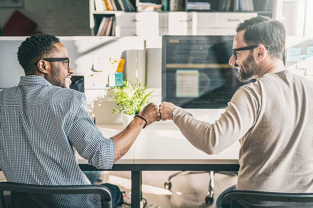 Two colleagues fist bumping while working at their desks in a modern office.